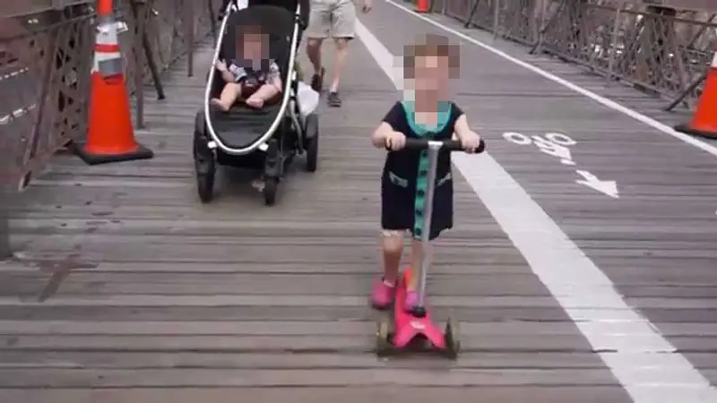 This is a Screenshot from a YouTube Video - A 2-Year-Old Rides her Scooter Along Brooklyn Bridge Without a Helmet (towards oncoming crowds) !!